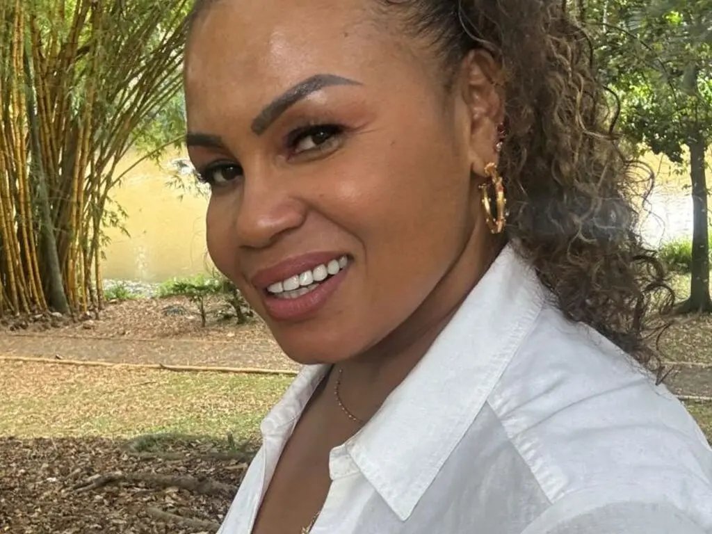 Smiling woman in a white button-down shirt posing outdoors in a lush garden setting.