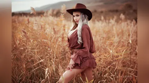 Blonde woman in a brown bohemian dress and wide-brimmed hat standing in a golden tall grass field at sunset.