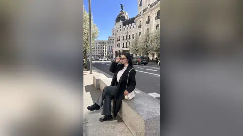 NashiroSakurai sits on a stone ledge wearing a black coat, white shirt, and dark trousers while wearing sunglasses on a city street.