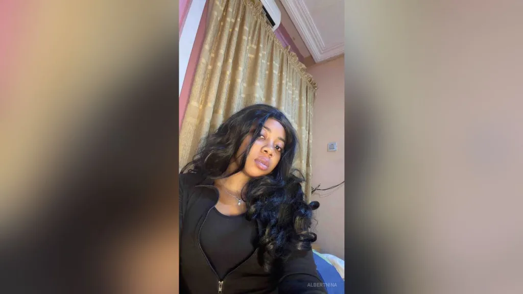AlbertNina — AlbertNina with long wavy black hair poses in a black top against gold curtains.