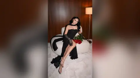 Elegant woman in a black evening gown and gloves sitting on a white chair with red roses in a moody setting.