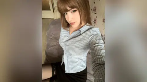 SharikaGlidden poses in a light blue pinstriped button-down shirt and black skirt, looking at the camera with long brown hair and bangs.