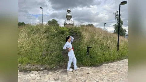 Woman in light blue lounge set posing on a stone path near a modern silver sphere sculpture under a cloudy sky.