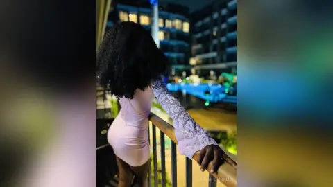 Woman in a pink lace dress standing on a balcony overlooking city lights at night.