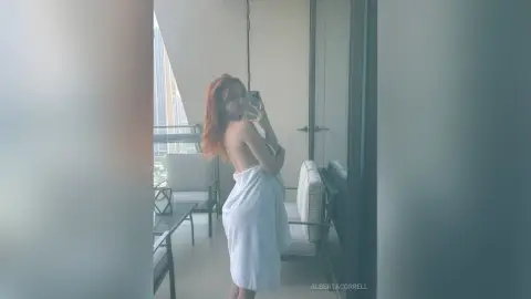 AlbertaCorrell poses in a white towel on a high-rise balcony, looking into her phone camera.