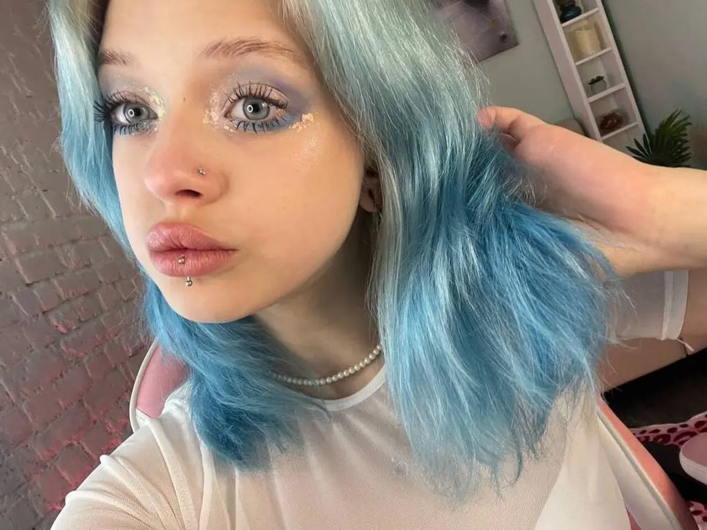 ClaudiaMaysons poses with her blue hair and heavy eye makeup, wearing a white mesh top and a pearl necklace.