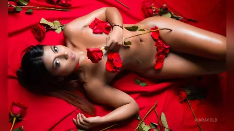 Sensual woman lying on red fabric surrounded by red roses in a romantic, moody setting.