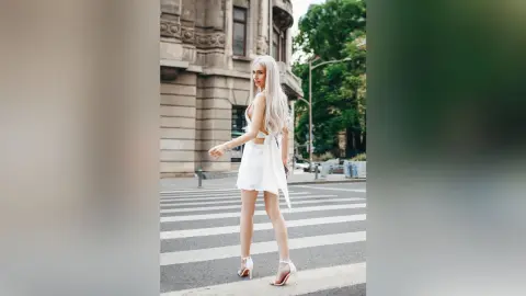Elegant blonde woman in a white cocktail dress walking across a city street near classical architecture.
