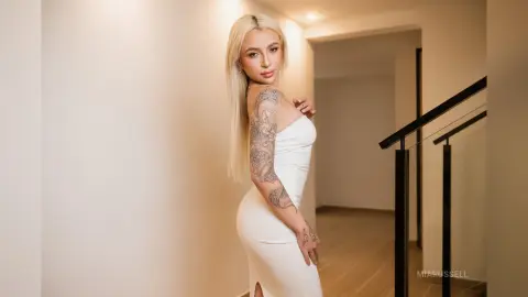 MiaRussell is a blonde woman with tattoos wearing a tight white dress, posing in a hallway.