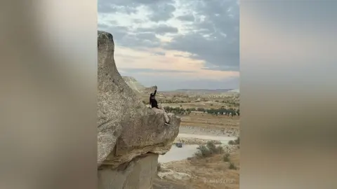 AlanaPearce sits on the edge of a steep, rocky cliffside under a cloudy sky, wearing a black top and light-colored pants.