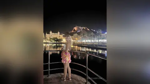 KaylaBryan stands on a balcony overlooking a city waterfront at night, wearing a white crop top and pink floral shorts.