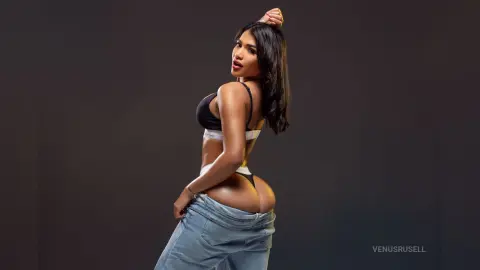 Confident woman in black lingerie and denim jeans posing against a dark studio background.