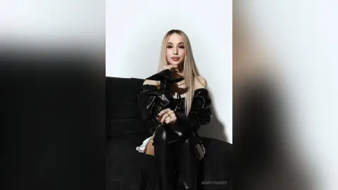 MarryDavies poses in black leather or vinyl clothing against a white background, resting her chin on her hand.