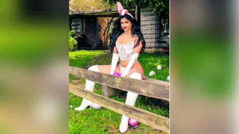 Smiling woman in white bunny costume and thigh-high boots sitting on a wooden fence in a lush green garden.