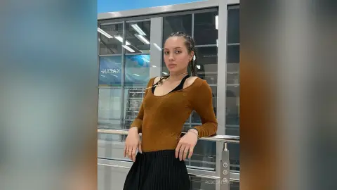 Young woman in a brown long-sleeve top and black skirt posing confidently in a modern glass building hallway.