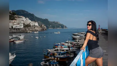 MissyJolie poses by a blue railing overlooking a harbor filled with boats, wearing a black tank top and striped shorts with sunglasses.