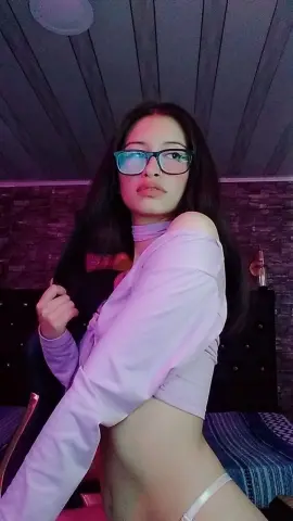 Young woman in a lavender long-sleeve crop top and glasses posing in a moody, dimly lit room.