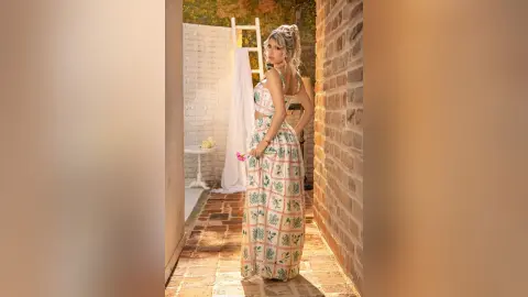 Blonde woman in a white floral maxi dress standing in a sunlit brick courtyard holding a pink flower.