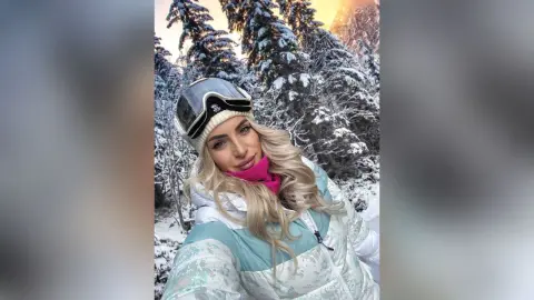 JessycaPage smiles in a white winter jacket and beanie, posing outdoors in a snowy forest.