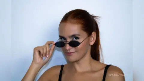 AmelieLautrec wears black sunglasses and a black tank top, posing with her hair pulled back in a ponytail against a plain white background.