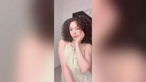 Smiling young woman with curly hair wearing a light green satin slip dress in a bright indoor setting.