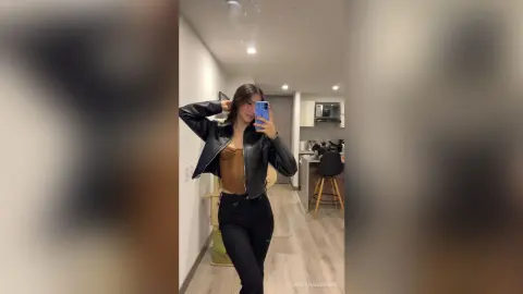 Young woman in a black leather jacket and brown top posing in a modern apartment hallway.