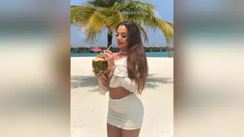 Young woman in white summer outfit sipping coconut water on a sunny tropical beach with palm trees.