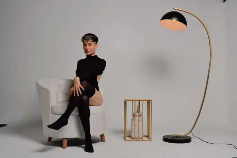 Elegant woman in a black bodysuit and tights sitting in a white armchair next to a modern gold floor lamp.