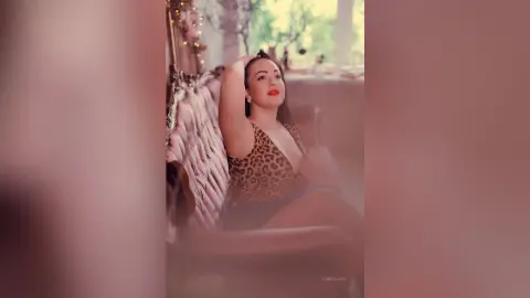 TannySmith with long dark hair and red lipstick poses in a leopard print top while sitting on a couch.