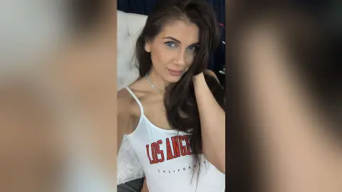 Young woman in a white Los Angeles graphic tank top relaxing in a bright indoor setting.