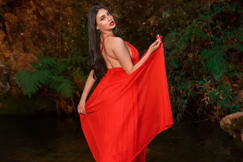 Elegant woman in a flowing red maxi dress posing by a lush jungle pond in a dreamy outdoor setting.