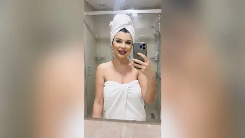 CapriAndersen poses in a bathroom mirror wearing a white towel wrapped around her chest and a matching white towel turban on her head.