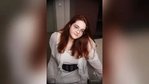 Redheaded woman in a white blouse and black belt smiling in a cozy indoor setting.