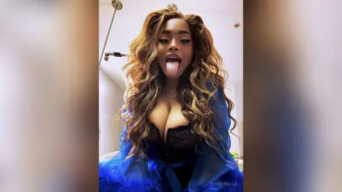BossyBella — BossyBella sticks her tongue out while wearing a blue lace bra and a blue feathered robe with long, wavy hair.