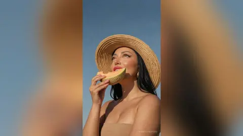 AnneThompson poses shirtless in a straw hat, holding a slice of cantaloupe near her mouth against a clear blue sky.