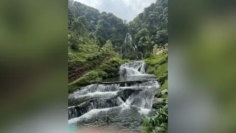 Serene tiered waterfall cascading through a lush green tropical forest under a cloudy sky.