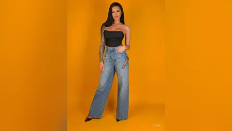 Young woman in a black tube top and wide-leg blue jeans posing against a vibrant yellow studio background.