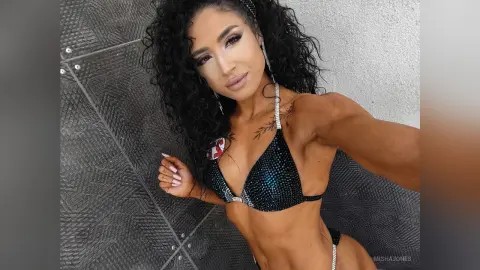 Fit woman with curly hair in a sparkling black bikini posing confidently against a textured gray wall.