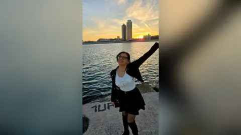 Smiling woman in a black jacket and white top enjoying a sunset view by the city waterfront.