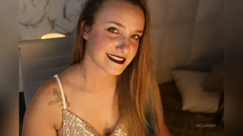 Smiling young woman in a sparkly gold sequin top posing in a cozy, dimly lit bedroom setting.