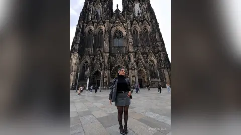 FabianaLima stands in front of a large gothic cathedral wearing a black turtleneck, a grey skirt, and black tights.