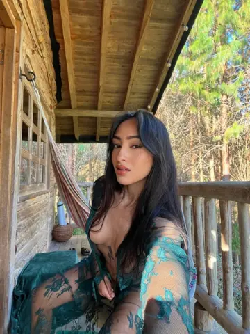 Young woman in a sheer green floral lace outfit relaxing on a wooden cabin porch in a sunny forest setting.