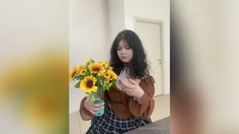 Young woman in a brown blouse holding a bright sunflower bouquet in a minimalist white room.