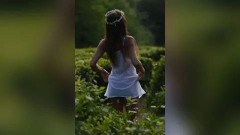 Dreamy woman in a white summer dress and flower crown walking through a lush green field at sunset.