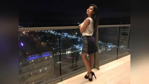 Elegant woman in a white sleeveless top and navy skirt posing on a city balcony at night.