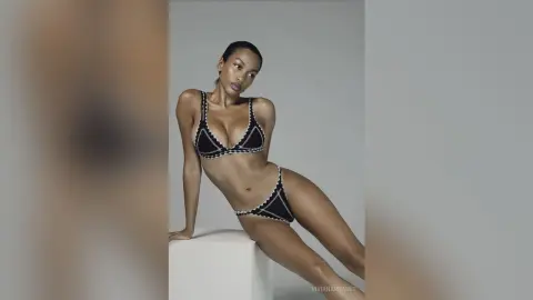 Athletic woman in a black patterned bikini posing confidently against a minimalist grey studio background.