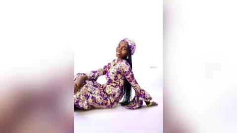 Smiling woman in a purple and gold floral print traditional dress sitting against a white background.