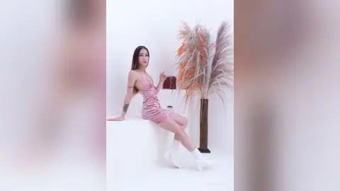 Young woman in a pink satin mini dress and white boots sitting in a bright minimalist studio with pampas grass.