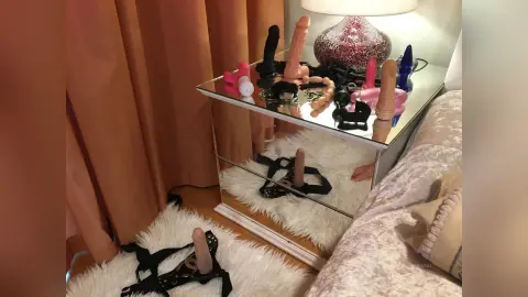 MilenaBia's collection of dildos and toys or black lace lingerie rests on a mirrored nightstand next to a bed.