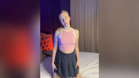 Young woman in a pink halter crop top and pleated navy skirt sitting on a bed in a dimly lit room.